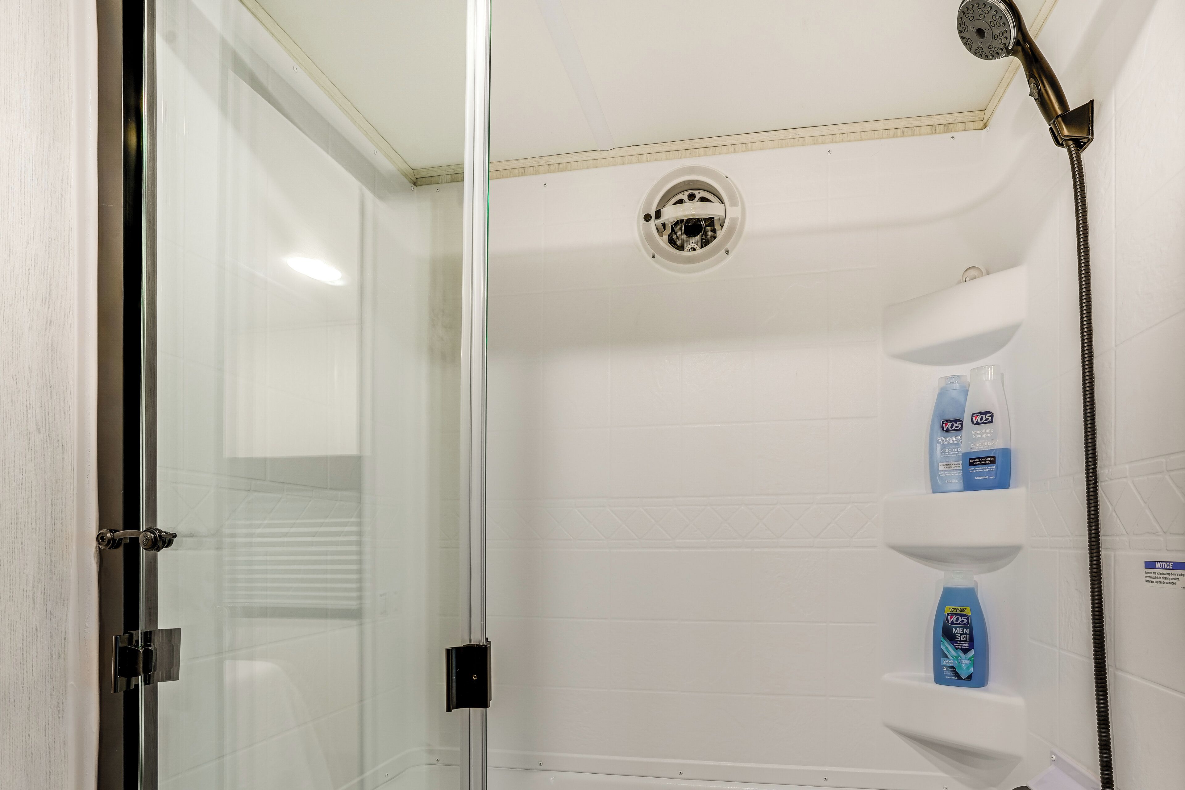 Doccia con porta in vetro, soffione e due flaconi di bagnoschiuma VOS su uno scaffale.