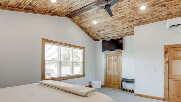A bedroom with a wooden ceiling, a bed with a beige bedspread, a window, a door, and a wall-mounted TV.
