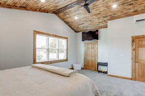 Quarto com teto de madeira, uma cama com colcha bege, janela, porta e televisão fixada na parede.