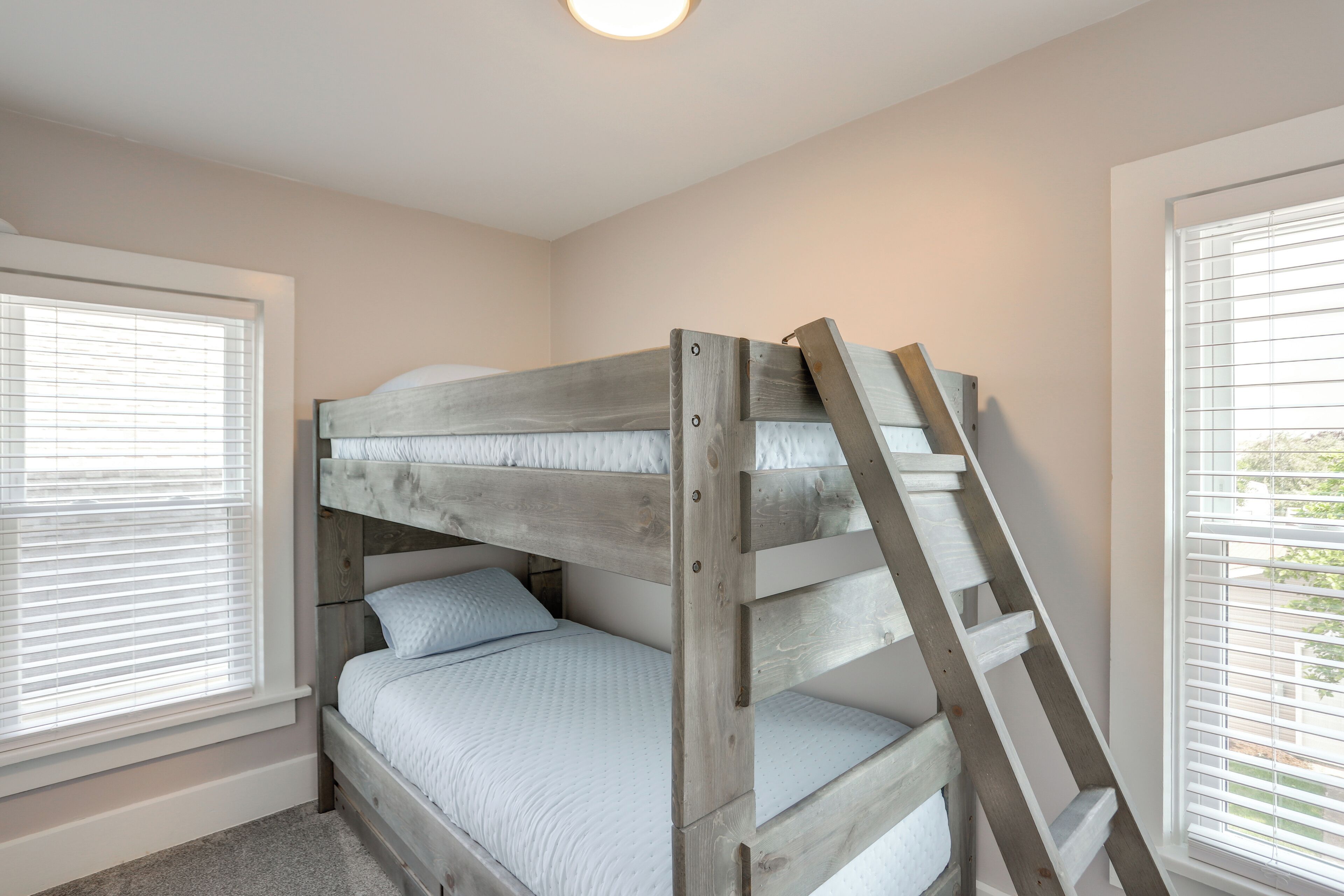 Een stapelbed met een houten ladder, wit beddengoed en ramen met zonwering.