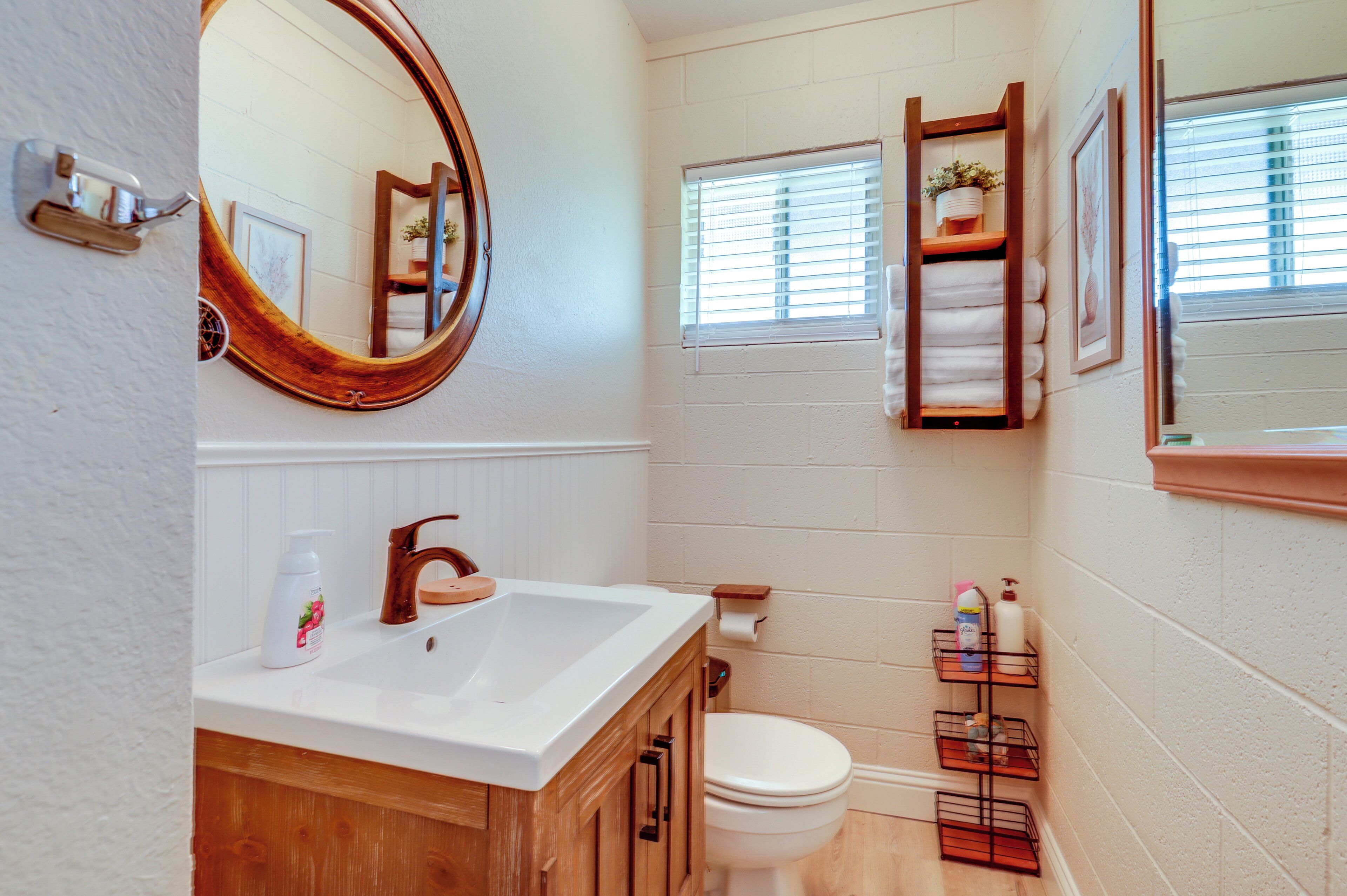 Une salle de bain avec un meuble-lavabo en bois, un lavabo blanc et des toilettes. Un miroir est placé au-dessus du lavabo et il y a une fenêtre avec des stores.