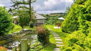 A garden pathway leading to a small structure with a bench and a table, surrounded by lush greenery and a pond. - Beautiful  villa for 2 people with WIFI (Leedstown)