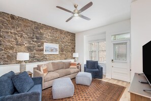 Una sala de estar con pared de piedra, ventilador de techo, sofá, otomanas, un sillón azul y televisor.