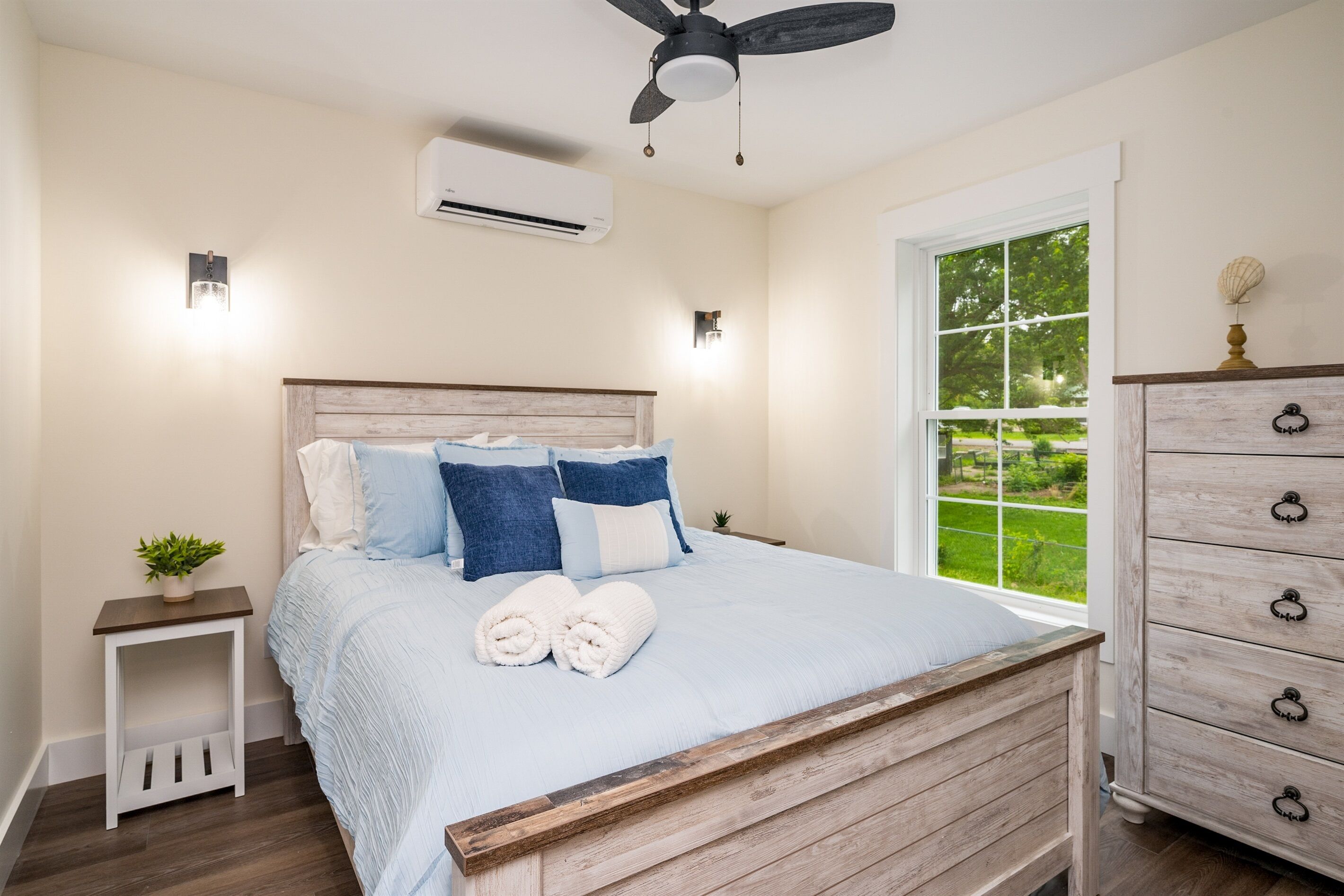 Ein Schlafzimmer mit einem hölzernen Bett, weißer Bettwäsche, einem Nachttisch mit Pflanze, einer Kommode mit Schubladen und einem Fenster mit Blick ins Grüne.