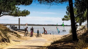 A sandy beach with bicycles and people, a water body in the background, and trees framing the scene.