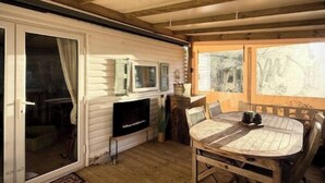 A cozy wooden deck with a round table and chairs, a television, and a view of trees through the windows.