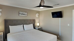 A bedroom with a large bed, a ceiling fan, a TV mounted on the wall, and a bedside table with a lamp. - Coconut’s Beach House | 2BD/1.5BA | Steps to Beach,  Lake View + Outdoor Shower (Pensacola Beach)
