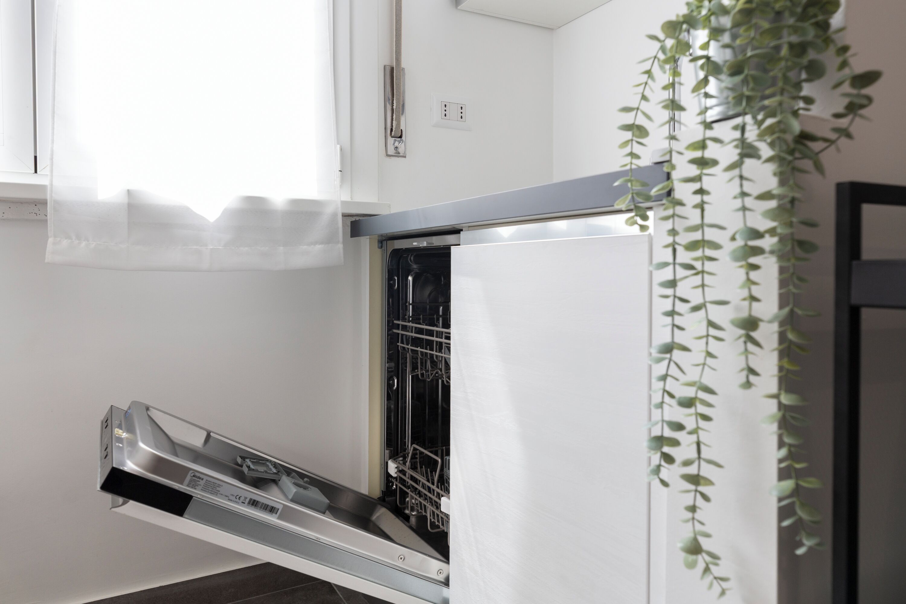 A kitchen with a dishwasher, a window with sheer curtains, and a hanging plant.