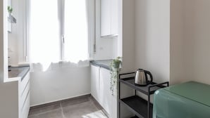 A compact kitchen with white cabinets, a window with sheer curtains, a small plant, a kettle on a black shelf, and a green suitcase. - notaMi - Fiordalisi 1 - Flower District (Milan)