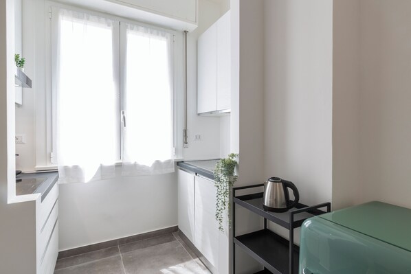 A compact kitchen with white cabinets, a window with sheer curtains, a small plant, a kettle on a black shelf, and a green suitcase.