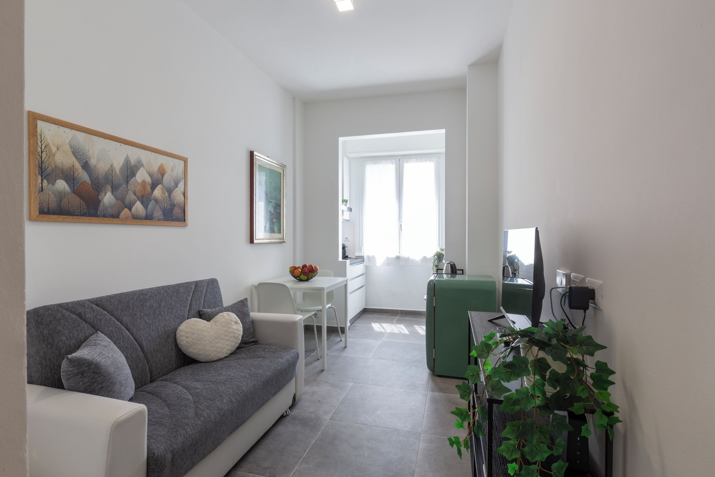 A modern living room with a grey sofa, a white dining table, and a green side table. There is a painting on the wall and a plant in the corner.