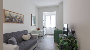 Una sala moderna con un sofá gris, una mesa de comedor blanca y una mesita auxiliar verde. Hay un cuadro en la pared y una planta en la esquina.