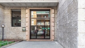A building entrance with a glass door, a small outdoor area with plants, and a metal pole. - notaMi - Fiordalisi 1 - Flower District (Milan)