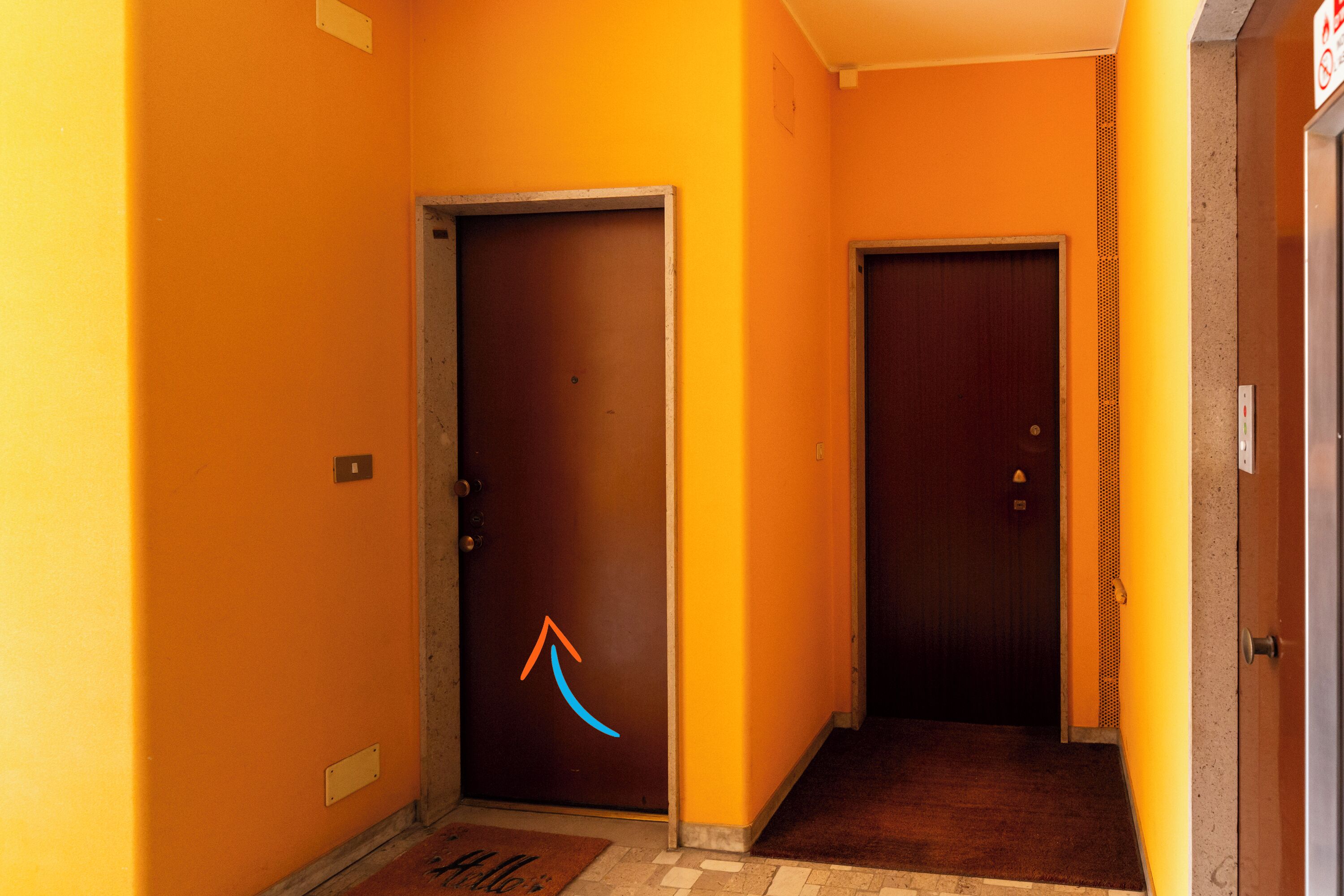 Um corredor com duas portas, uma com uma seta azul apontando para cima e um tapete de entrada com a palavra 'Hello'.