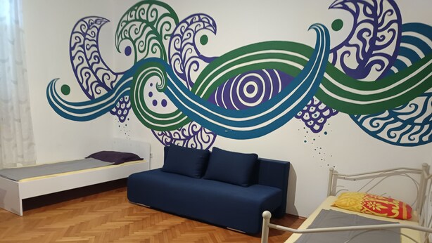 Une pièce avec un canapé bleu, un lit recouvert d’une couverture à motifs jaunes et rouges, et un mur orné d’œuvres d’art abstraites.