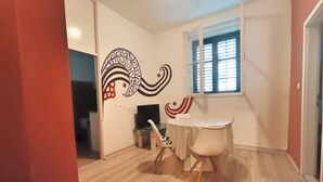 Une pièce avec un plancher en bois, une table blanche avec une chaise, un téléviseur posé sur un meuble et une fenêtre avec des stores.