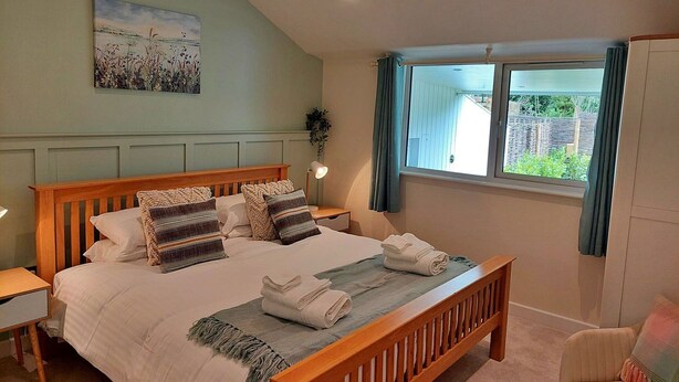 A bedroom with a wooden bed, a painting on the wall, a window with curtains, and a view of greenery outside. - Stunning  villa for 2 guests with hot tub, WIFI (Kingsteignton)