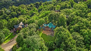 A large, multi-story wooden house surrounded by dense forest, with a well-maintained lawn and a winding driveway.