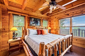 Ein Schlafzimmer in einer Blockhütte mit einem Bett, zwei Nachttischen, einem Deckenventilator und großen Fenstern mit Jalousien.