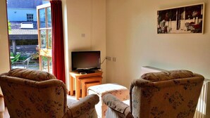 A living room with two floral armchairs, a television on a wooden stand, and a large window with a red curtain.