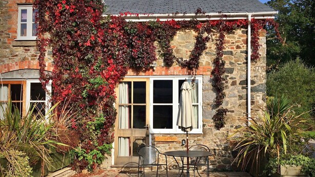 A stone cottage with ivy-covered walls, a patio with a table and chairs, and a sign reading "Virginia Cottage."