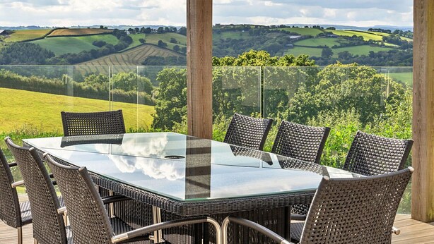 Une table à plateau en verre, entourée de chaises en osier, installée sur une terrasse en bois offrant une vue sur des collines ondoyantes.