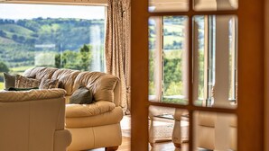 Un salon avec un canapé beige, des coussins à motifs et une vue sur un paysage verdoyant grâce à de grandes fenêtres.