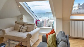 Quarto de loft acolhedor com teto inclinado, um amplo claraboia, vista para o mar, uma cama e um espaço de estar com sofá e poltrona.