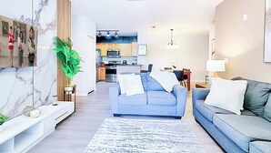 A modern living room with a sofa, a coffee table, and a dining area with a table and chairs. There is a kitchen in the background with cabinets and appliances. - 8908 Family Fun in Kissimmee Spacious 4-bedroom Apartment Near Theme Parks (Kissimmee)