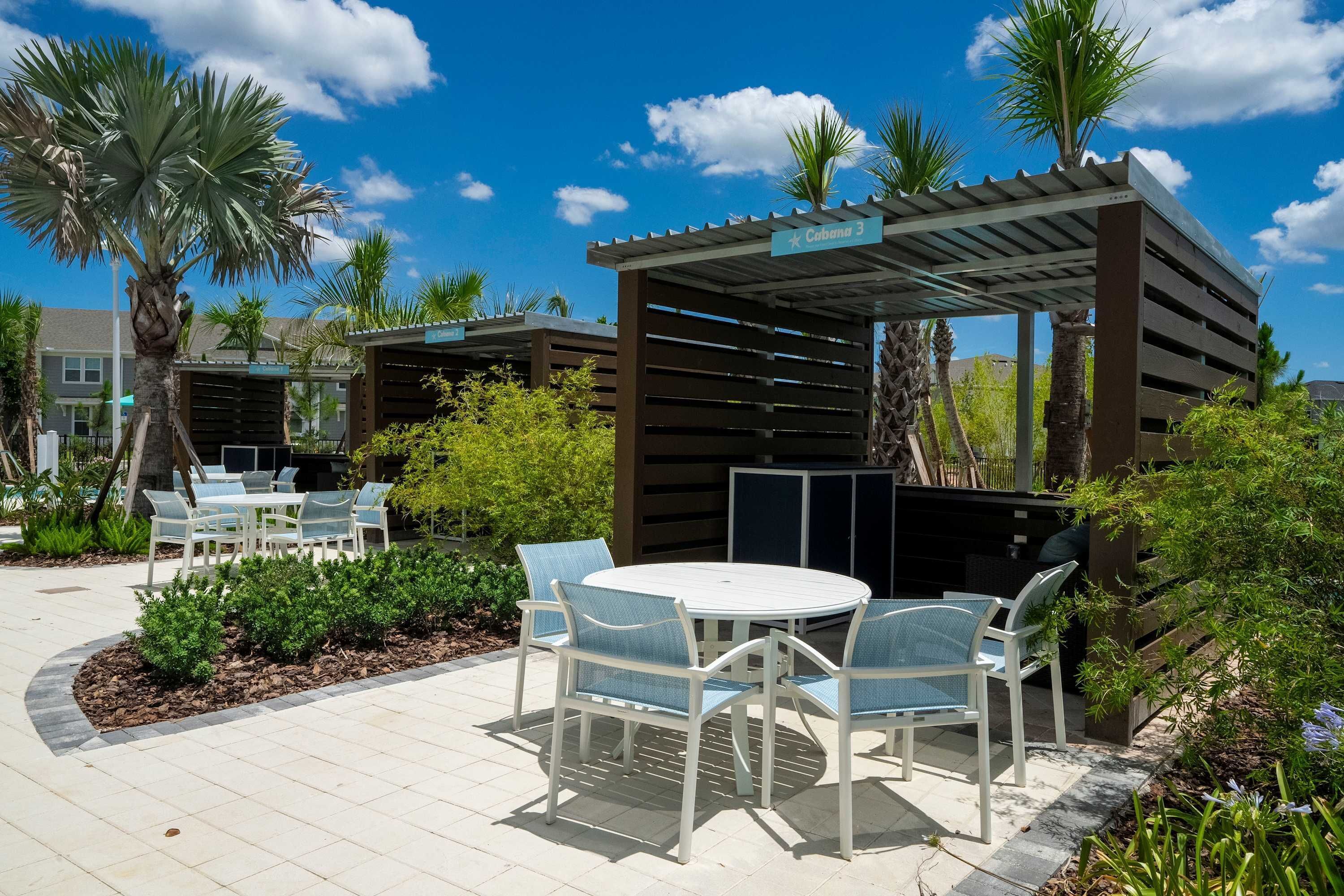 An outdoor patio area with a round table and chairs, surrounded by palm trees and a pergola structure.