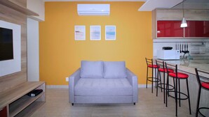 A living room with a grey sofa, a TV on the left, and a kitchen area with red chairs and a white microwave.