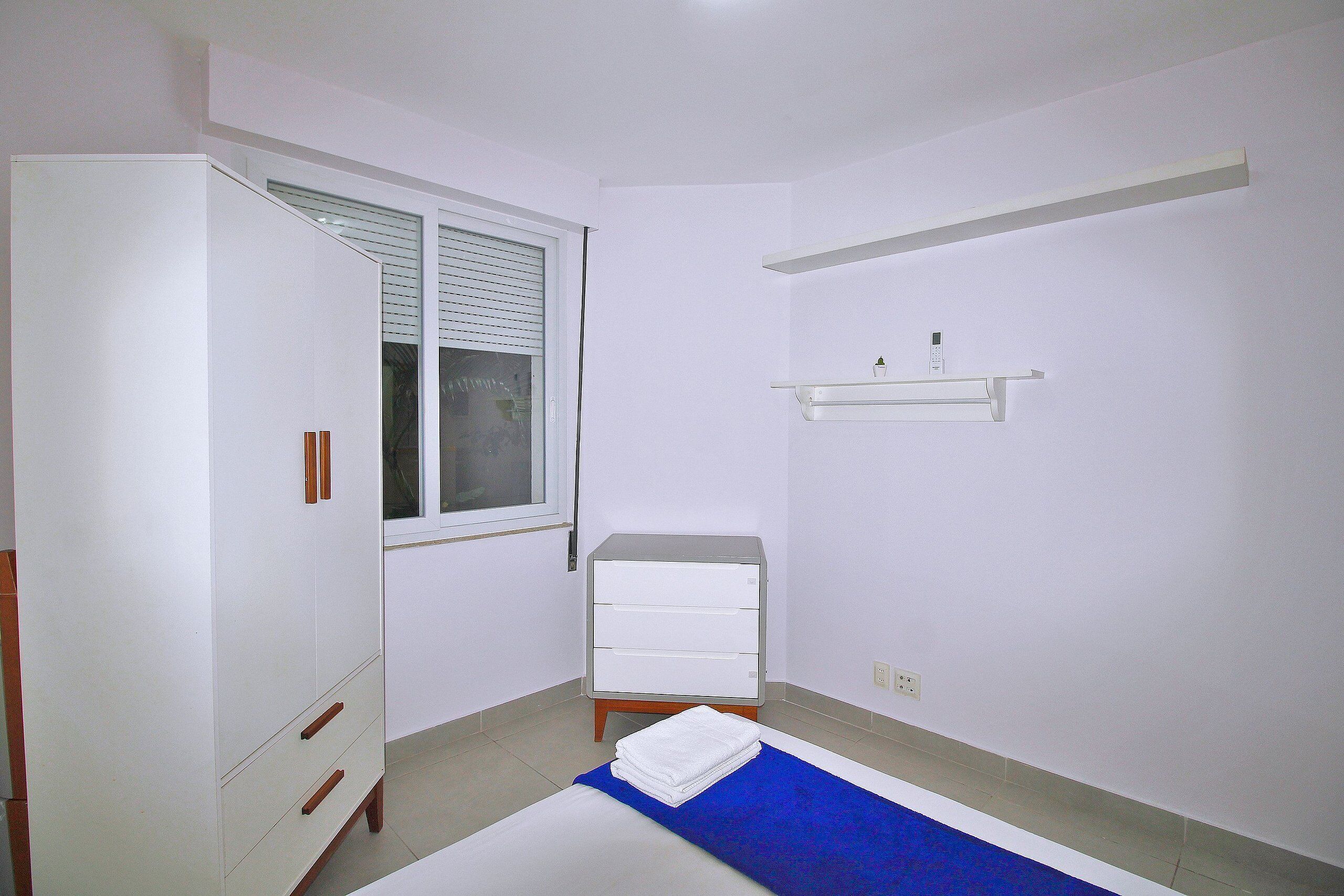 A bedroom with a bed, a wardrobe, a small bedside table, a window with blinds, and a wall-mounted shelf.