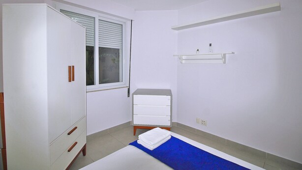 A bedroom with a bed, a wardrobe, a small bedside table, a window with blinds, and a wall-mounted shelf. - Conforto e Estilo em Copacabana D056 (Rio de Janeiro)