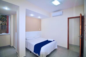 A single-bed room with a white bed, a blue blanket, a window with a red curtain, and an air conditioning unit on the wall. - Conforto e Estilo em Copacabana D056 (Rio de Janeiro)