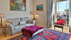 A modern living room with a grey sofa, a red ottoman, and a patterned rug. There is a painting of a cityscape on the wall, a lamp on a side table, and a view of the city through large windows.