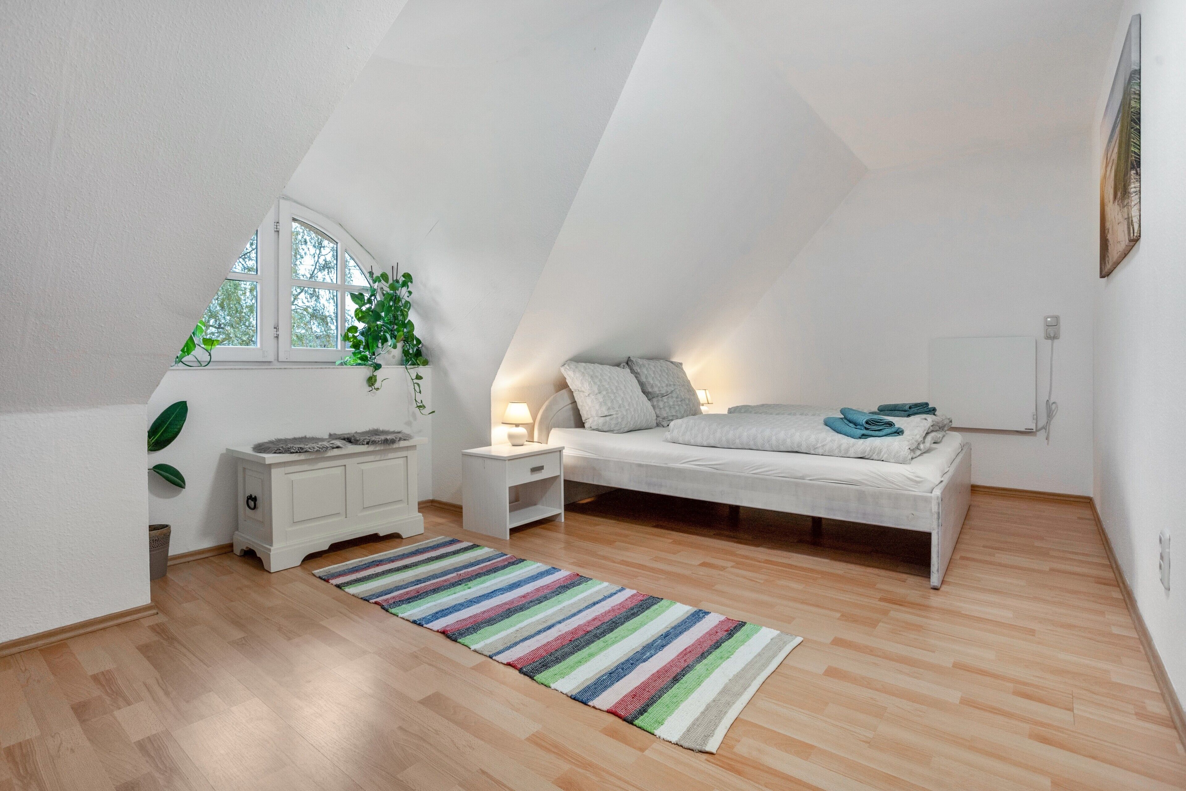Ein Schlafzimmer mit schrägen Wänden, einem Bett, Nachttischen, einem Fenster mit Blick und einem Streifenmuster-Teppich.