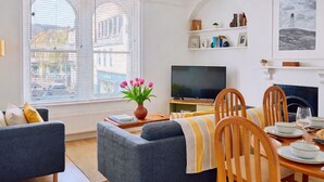 Een woonkamer met een bank, televisie, eettafel met stoelen en een open haard.