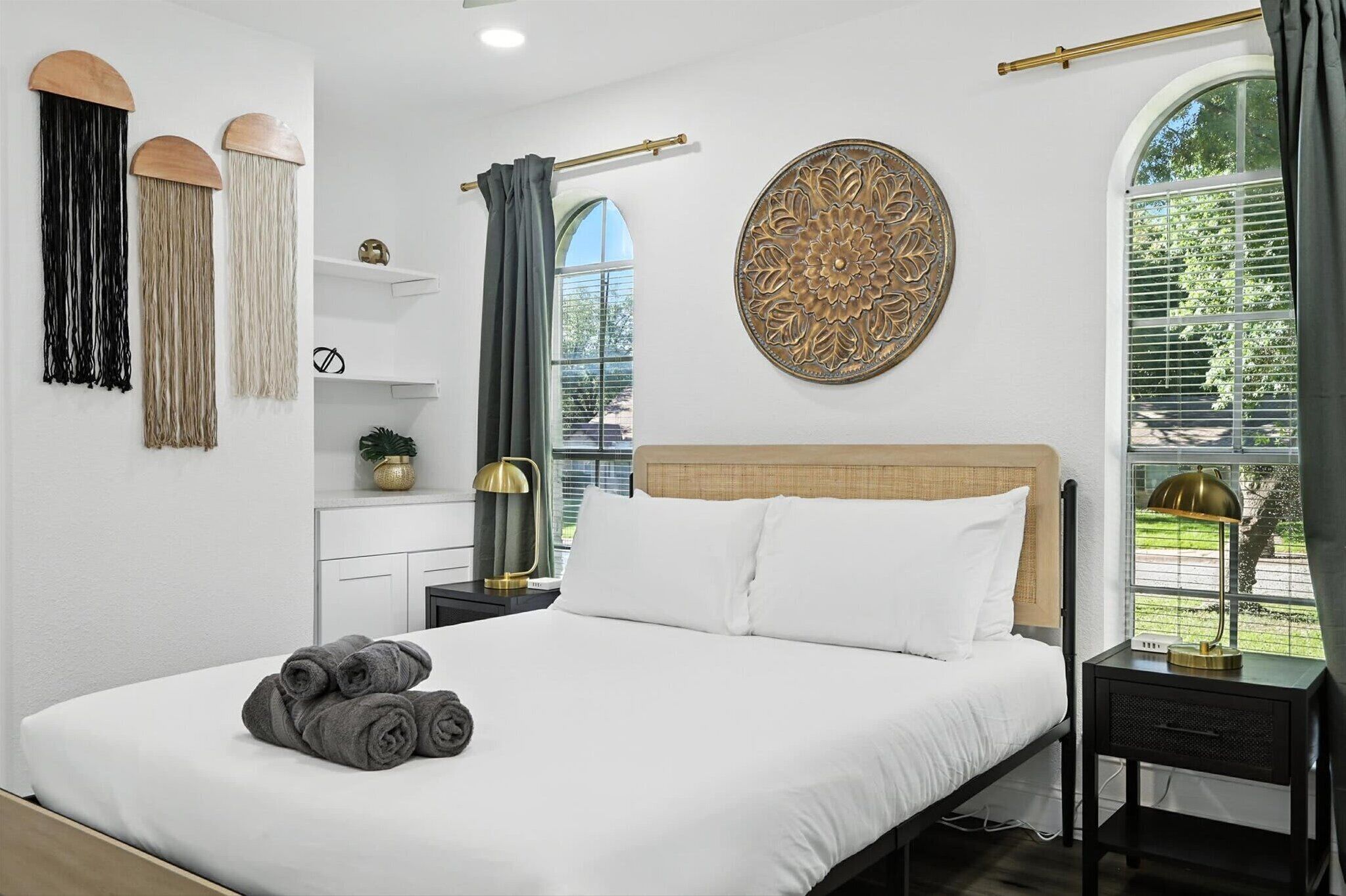 A neatly made bed with white linens and rolled towels, a bedside table with a lamp, and a decorative wall hanging.