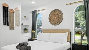 A neatly made bed with white linens and rolled towels, a bedside table with a lamp, and a decorative wall hanging.