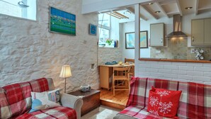 A cozy living room with a red plaid sofa, a wooden coffee table, and a lamp. There is a dining area with a wooden table and chairs. The room has a stone wall and a large window. - Beautiful  villa for 4 people with WIFI and pets allowed (Cawsand)