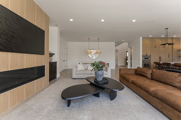 A modern living room with a flat-screen TV, a sofa, a coffee table, and a dining area with a kitchen in the background.