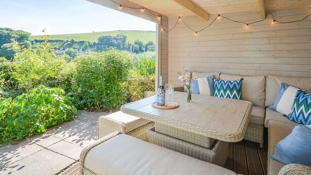 An outdoor patio with wicker furniture, a table with a bottle and glasses, and string lights. - Beautiful  villa for 8 people with WIFI and pets allowed (Cawsand)