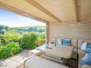Une terrasse extérieure avec des meubles en osier, une table avec une bouteille et des verres, et des guirlandes lumineuses.