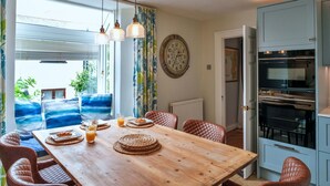 A dining area with a wooden table, woven placemats, and leather chairs. A large window with curtains allows natural light to enter. - Beautiful  villa for 8 people with WIFI and pets allowed (Cawsand)