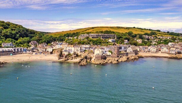 A coastal town with a historic stone building near the water, surrounded by residential houses and a hilly landscape.