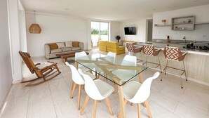 A modern living space with a glass dining table surrounded by white chairs, a sofa, and a yellow armchair. A kitchen with granite countertops and a TV mounted on the wall is visible. - Apartment for 8 Guests in Sabanilla a Few Steps from the Sea (Sabanilla)