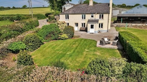 Une maison à deux étages avec une pelouse et un jardin bien entretenus, un espace patio avec des sièges d’extérieur et un grand arbre en arrière-plan.