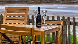 A wooden table with a bottle of wine and two glasses on a wooden chair, set against a wooden fence with a beach and ocean in the background.