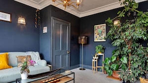 A modern living room with a dark blue wall, a white sofa with yellow cushions, a glass coffee table, and various potted plants.