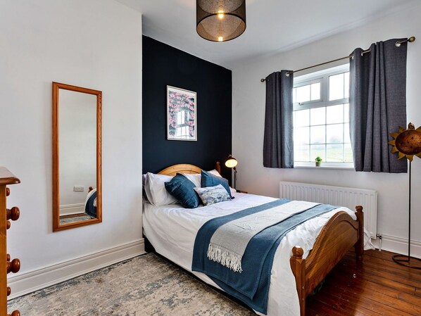 A bedroom with a bed, a bedside table, a mirror, a window with curtains, and a radiator.